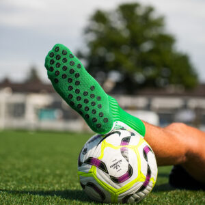 Groene Bridge gripsok met zichtbaar antislip-noppenpatroon; voet rust op witte voetbal op kunstgras.