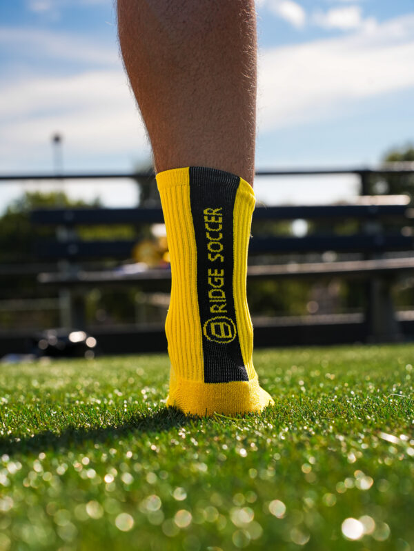 Gele Bridge gripsok met zwarte verticale baan en gele tekst “BRIDGE SOCCER” op kunstgras, close-up van achteren.