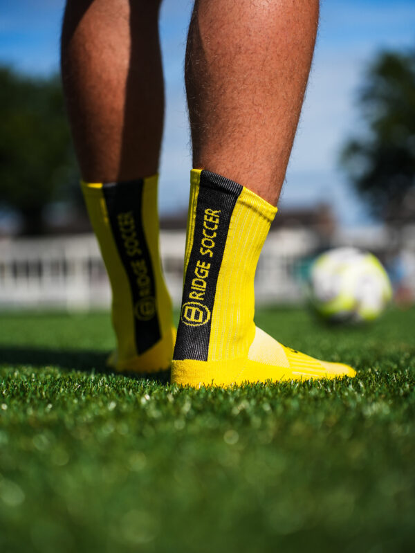 Gele Bridge gripsokken met zwarte verticale baan en “BRIDGE SOCCER”; close-up op kunstgras, voetbal onscherp op achtergrond.