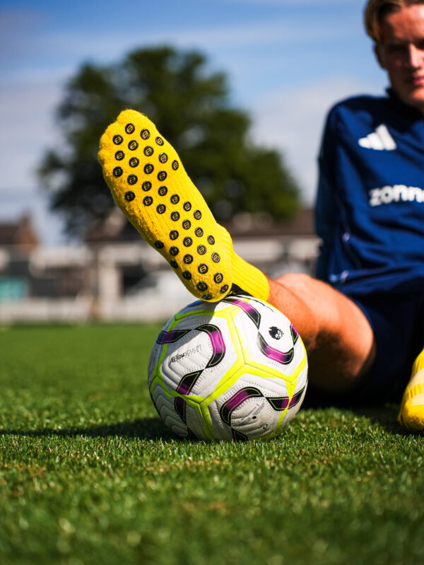 Gele Bridge gripsok met zwarte antislip-noppen; voet rust op witte voetbal op gras.