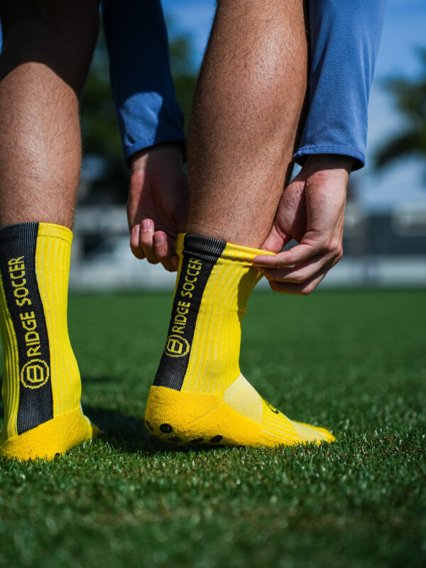 Speler trekt gele Bridge gripsok met zwarte baan omhoog; handen bij enkel, op grasveld.