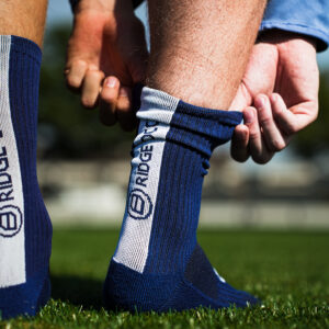 Speler trekt donkerblauwe Bridge gripsok met witte verticale baan omhoog; handen bij enkel op grasveld.