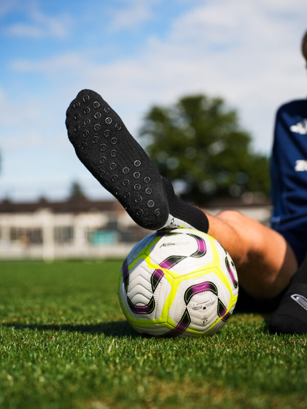 Zwarte Bridge gripsok met witte antislip-noppen; voet rust op witte voetbal op gras.
