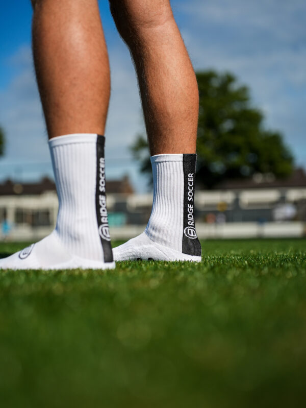 Witte Bridge gripsokken op gras; close-up van opzij/achteren met zwarte verticale baan en “BRIDGE SOCCER”.