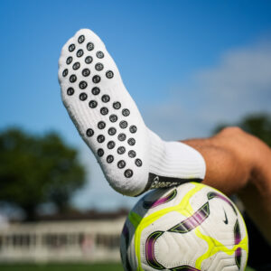 Witte Bridge gripsok met zwarte antislip-noppen; voet rust op witte voetbal op gras, blauwe lucht op achtergrond.